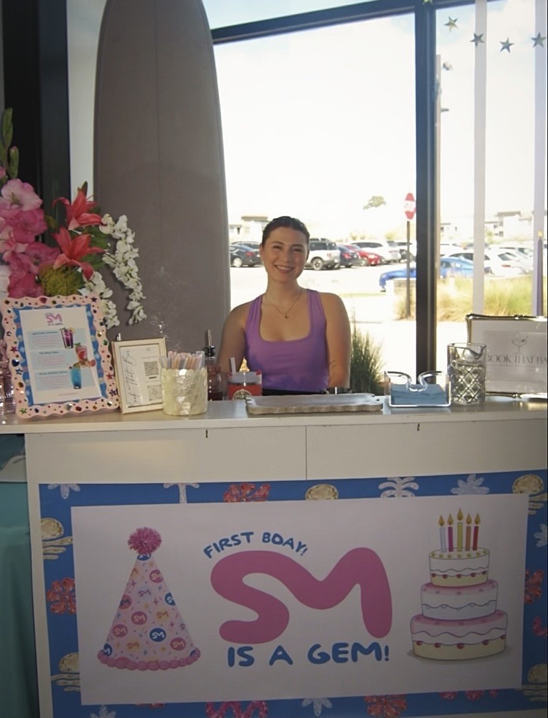 Bartender in purple behind a birthday bar with flowers and a custom sign