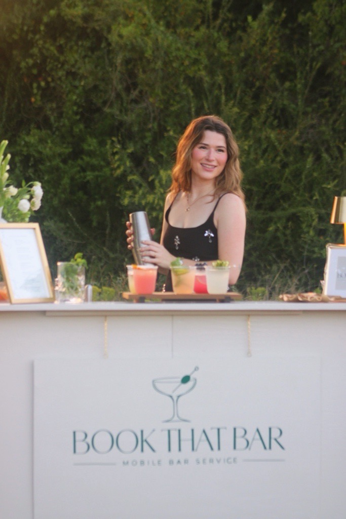 Morgan smiling behind the Book That Bar mobile bar with a cocktail shaker