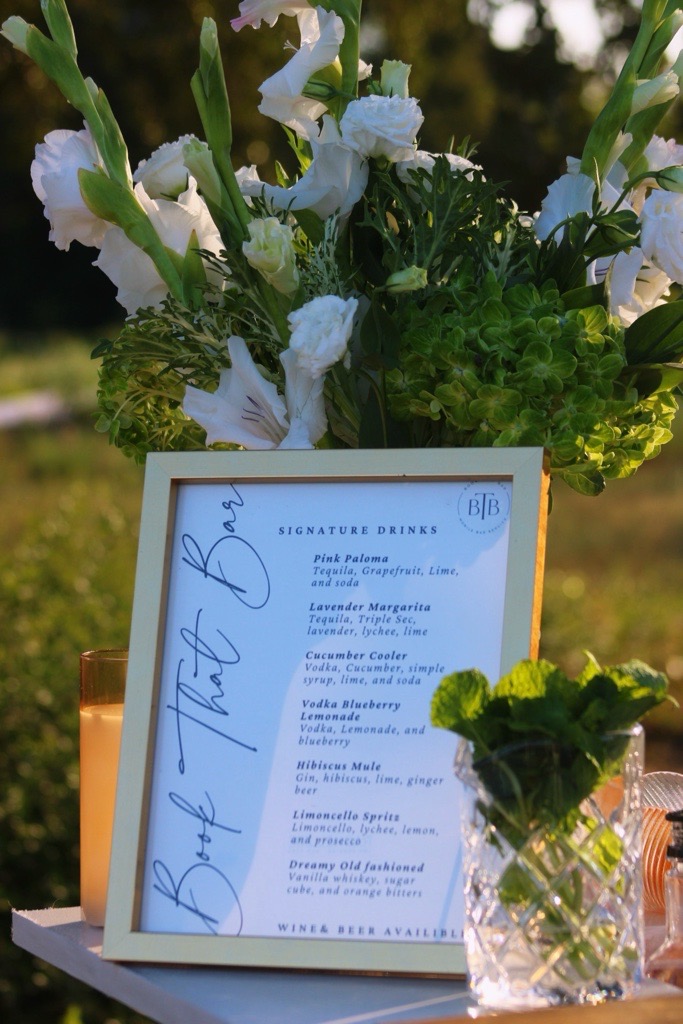 A framed Book That Bar signature drink menu surrounded by white flowers and fresh mint